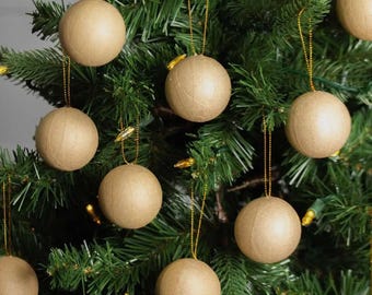 May include: A collection of spherical ornaments made from light brown, textured paper material. Each round ornament is suspended by a thin gold string and hangs from the green branches of a Christmas tree, creating a uniform, neutral display.