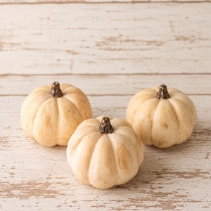 May include: Three small, round, cream-colored decorative pumpkins with visible vertical ridges and dark brown stems. Each pumpkin features a smooth, matte finish with subtle light brown shading in the crevices, arranged in a triangular formation.