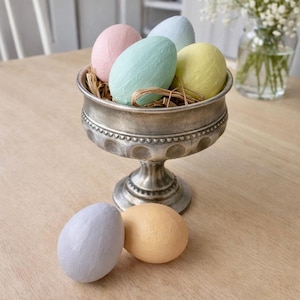 May include: A silver pedestal bowl filled with pastel-colored decorative eggs. The eggs are pink, blue, green, and yellow, nestled in straw. Two additional eggs, one lavender and one peach, rest beside the bowl on a wooden surface. A vase of white flowers is in the background.