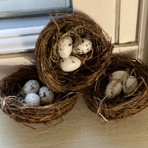 May include: Three woven nests, each filled with speckled white eggs. The nests are made of dried twigs and are arranged in a cluster. The eggs have brown speckles, and the nests are a natural brown color.