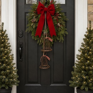 May include: A green pine wreath featuring a large red velvet bow, pinecones, and red berries, with two hanging wire bells decorated with small red and white checkered ribbons, all mounted on a black door between two small lit evergreen trees.