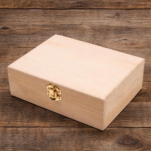 May include: A rectangular, unfinished wooden box with a gold-colored clasp. The box is made of light-colored wood and has a simple, clean design. The clasp is centered on the front of the box.