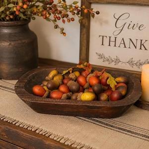May include: A dark brown wooden dough bowl filled with a collection of artificial acorns in shades of orange, yellow, and brown, accented with small faux autumn leaves. The bowl has a rustic, carved texture and an elongated, rectangular shape.
