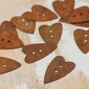 May include: A collection of heart-shaped buttons with two holes each. The buttons have a rustic, weathered appearance with a reddish-brown color. They are scattered on a white surface with visible wood grain.