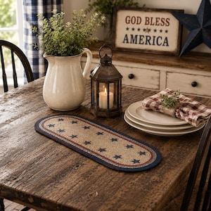 May include: A rustic wooden table is set with a patriotic theme. A beige oval rug with blue stars and a red border sits in the center. A white pitcher, lantern, and plates are also on the table. A sign reads "GOD BLESS AMERICA."