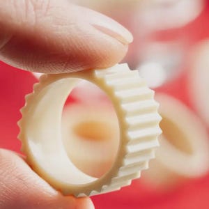 May include: A cream-colored, ring-shaped napkin holder with a ridged exterior. The napkin ring is held between fingers against a blurred red background. The ring's design features a series of vertical ridges around its circumference.