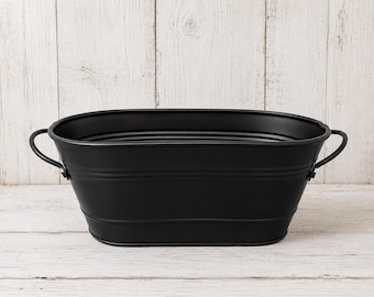 May include: An oval-shaped black metal tub featuring a smooth finish with horizontal ribbed detailing along the sides. The container includes two curved metal handles attached to each side with rivets, providing a functional design for storage.