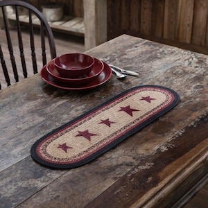 May include: Oval jute table runner with burgundy stars and a black border. The runner is placed on a rustic wooden table, alongside burgundy-colored dinnerware and silverware. The runner measures approximately 36 inches long.