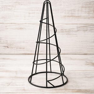 May include: A black metal cone-shaped decorative object. The structure features a circular base, vertical supports, and a spiral design wrapping around the cone. The object is set against a light wood-grain background.