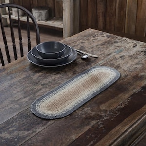 May include: An oval-shaped, braided table runner in shades of gray, beige, and cream. The runner is placed on a rustic wooden table, with a dark gray bowl and plates, and silverware visible in the background. The table setting suggests a dining theme.