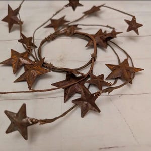 May include: A circular wreath constructed from thin, brown, twig-like wire adorned with multiple five-pointed metal stars. The stars feature a weathered, rusted brown finish and are attached at various intervals along the spiraling wire frame.