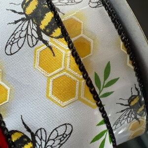 May include: White ribbon with a repeating pattern of black and yellow bumblebees, yellow honeycomb shapes, and green leaves. The ribbon's edge is trimmed with a black, textured border. The design is suitable for crafts and gift wrapping.