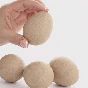 May include: A hand holds a light brown, egg-shaped object above three similar objects. The objects have a textured surface and are likely made of stone or a similar material. The background is plain white.