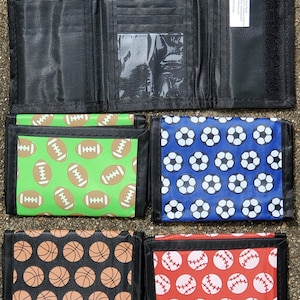 May include: Four tri-fold wallets with different sports-themed patterns. The wallets are black with a clear plastic window on the front. The patterns are footballs on a green background, soccer balls on a blue background, basketballs on a black background, and baseballs on a red background.