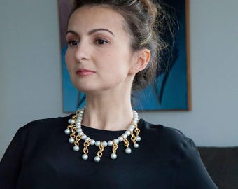 May include: A statement necklace featuring a thick gold-toned chain base adorned with dangling light grey spherical beads. The necklace is worn against a plain black top, showcasing a layered design where the beads hang from the gold chain links.