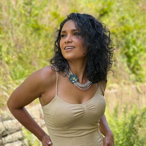 May include: A multi-strand necklace featuring small, light-colored beads arranged in several layers, accented by a prominent, dark teal, flower-shaped pendant with a central gold-toned detail, worn with a beige, sleeveless, ruched-front top.