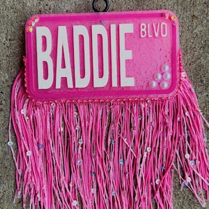 May include: A pink license plate-shaped accessory with the word "BADDIE" in white block letters and "BLVD" in smaller letters. The accessory has a fringe of pink cords and is embellished with sequins and small beads.
