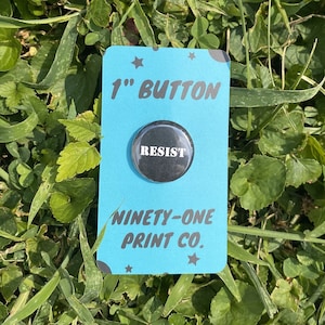 May include: A 1-inch button with the word "RESIST" in white text on a black background. The button is on a blue card with white stars and the text "1" inch button" and "Ninety-One Print Co."