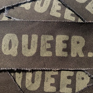 May include: A black fabric patch with the word "QUEER" printed in green letters.