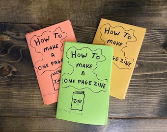 May include: Three colorful paperbacks titled "How To Make A One Page Zine" with illustrations of a book and the word "Zine" on the cover. The books are stacked on a wooden surface.