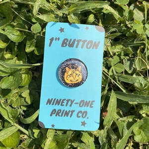 May include: A 1-inch button featuring a black and gold illustration of a cat with three eyes. The button is on a blue background with white stars and the text "Ninety-One Print Co."