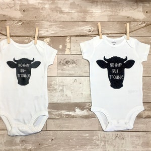 Peut inclure: Deux bodies blancs pour b&eacute;b&eacute; avec des silhouettes de vache noires et le texte "Nothin' But Trouble" imprim&eacute; dessus. Les bodies sont suspendus &agrave; une corde &agrave; linge avec des pinces &agrave; linge en bois.