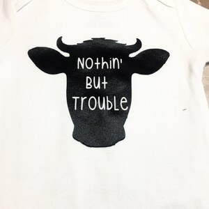 Peut inclure: Un body blanc pour b&eacute;b&eacute; avec une silhouette noire de t&ecirc;te de vache et le texte "Nothin' But Trouble" imprim&eacute; dessus.