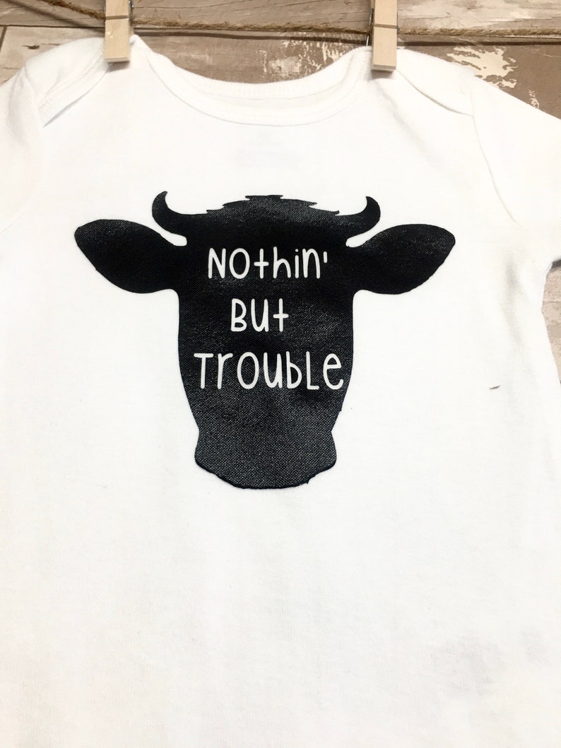 Peut inclure: Un body blanc pour b&eacute;b&eacute; avec une silhouette noire de t&ecirc;te de vache et le texte "Nothin' But Trouble" imprim&eacute; dessus.