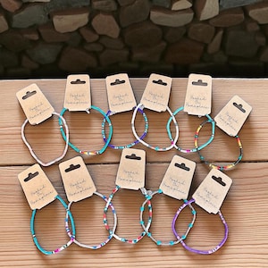 May include: A collection of beaded bracelets in various colors, including turquoise, white, and purple, displayed on a wooden surface. Each bracelet is attached to a brown tag that reads "Hooked On Homeplace". The bracelets feature colorful patterns.