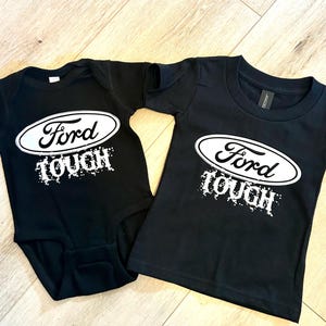 May include: Black baby onesie and t-shirt with the Ford logo in white inside an oval, and the word "TOUGH" in a distressed font. The baby onesie has short sleeves and the t-shirt has a crew neck.