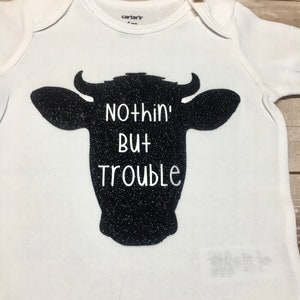 Peut inclure: Combinaison blanche pour b&eacute;b&eacute; avec une silhouette noire de t&ecirc;te de vache et le texte "Nothin' But Trouble".