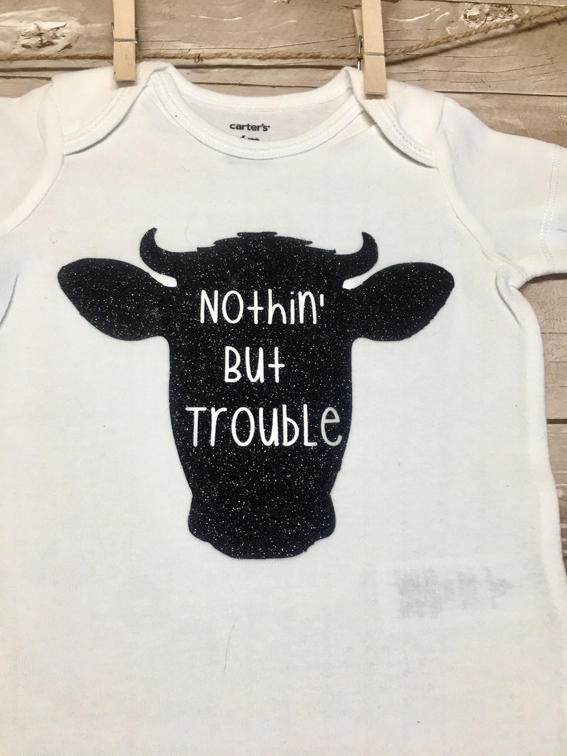 Peut inclure: Combinaison blanche pour b&eacute;b&eacute; avec une silhouette noire de t&ecirc;te de vache et le texte "Nothin' But Trouble".