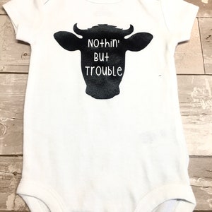 Peut inclure: Combinaison blanche pour b&eacute;b&eacute; avec une silhouette noire de t&ecirc;te de vache et le texte "Nothin' But Trouble".