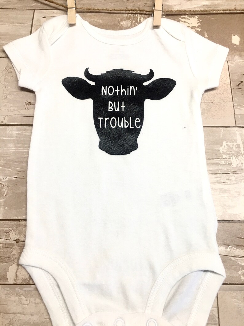 Peut inclure: Combinaison blanche pour b&eacute;b&eacute; avec une silhouette noire de t&ecirc;te de vache et le texte "Nothin' But Trouble".