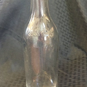 Peut inclure: Bouteille en verre transparent avec un corps arrondi et un col étroit. La bouteille porte des lettres en relief sur le devant et une marque circulaire sur le fond. La bouteille est vide et semble être vintage.