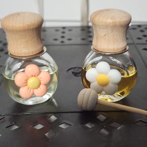 May include: Two small glass bottles with wooden stoppers, each containing a liquid and a decorative flower. One flower is peach and yellow, the other is white and yellow. A small wooden honey dipper rests nearby.