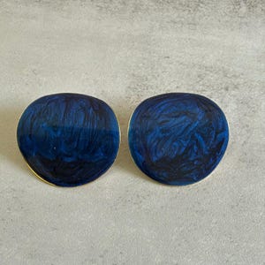 May include: A pair of round stud earrings featuring a glossy, deep blue marbled finish with swirling dark navy patterns and a thin gold-toned metal edge. The surface of each earring has a smooth, reflective appearance with irregular, organic shapes.