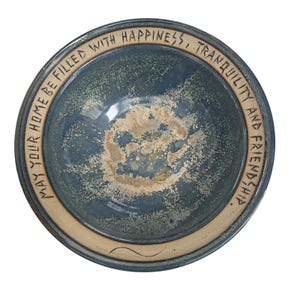 May include: A round ceramic bowl with a blue and grey glazed interior featuring a speckled beige center. The wide, light-colored rim is inscribed with the black text: MAY YOUR HOME BE FILLED WITH HAPPINESS, TRANQUILITY AND FRIENDSHIP.