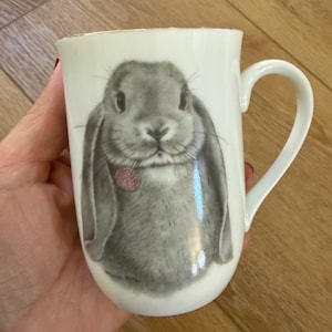 May include: A white ceramic mug featuring a detailed gray illustration of a lop-eared rabbit. The rabbit is depicted with long ears and a small pink raspberry near its mouth. The mug has a smooth, glossy finish and a curved handle on the right side.