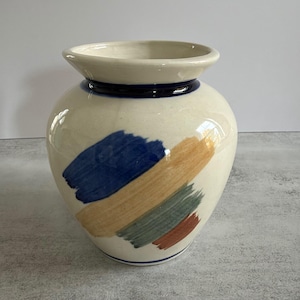 Peut inclure: Un vase en céramique de couleur crème avec un bord évasé et un corps arrondi. L'extérieur présente des coups de pinceau peints à la main en bleu, beige, vert sauge et terre cuite, avec une fine bande bleu foncé autour du col et de la base.