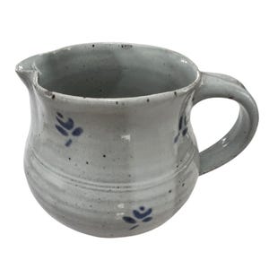 May include: A light gray ceramic creamer pitcher featuring a speckled finish and small, hand-painted dark blue floral motifs. The pitcher has a rounded body, a curved handle on the right side, and a small pouring spout at the top rim.