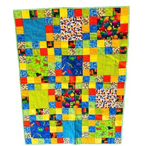 May include: A colorful patchwork quilt with a variety of patterned squares. The quilt features squares with dinosaurs, frogs, and other playful designs in shades of blue, green, red, and yellow. The quilt is bordered with a green fabric.