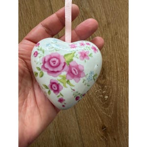May include: A heart-shaped ceramic ornament featuring a white base with a floral pattern of pink roses, green leaves, and small blue flowers. A pale pink ribbon is attached to the top for hanging, providing a decorative accent to the smooth surface.