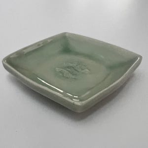 May include: A square ceramic dish with a glossy, pale sage green glaze. The center of the dish features a subtle, embossed relief design of two small fish swimming in opposite directions, creating a textured focal point on the smooth surface.