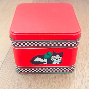 Puede incluir: Una lata de metal roja con forma cuadrada y tapa a juego. La lata presenta una ilustración de gato en blanco y negro con un lazo verde. El diseño incluye un patrón de tablero de ajedrez en blanco y negro.