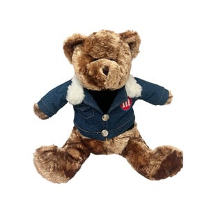 May include: A brown teddy bear wearing a denim jacket with a white faux fur collar. The jacket has two silver buttons and a red and white patch on the chest. The bear is sitting with its legs spread.
