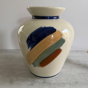 Puede incluir: Un jarrón de cerámica de color crema con un cuerpo ancho y redondeado y un borde acampanado. El exterior presenta una serie de pinceladas diagonales pintadas a mano en azul marino, beige, verde salvia y marrón terracota.