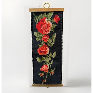Könnte beinhalten: Ein vertikaler Wandbehang in Stickoptik mit dunkelblauem Hintergrund und einem zentralen Blumenmuster aus roten und orangefarbenen Rosen sowie grünen Blättern. Das Textil wird von Holzleisten gehalten und besitzt einen goldenen Metallring.