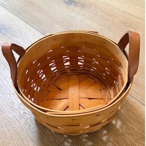 Peut inclure: Un panier rond en bois tressé avec une finition brun clair et deux poignées en cuir robustes fixées sur les côtés. Le panier est construit avec de fines bandes de bois superposées formant une base circulaire et des parois au tissage visible.