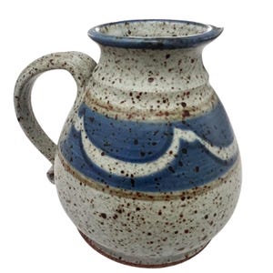 Peut inclure: Une cruche en céramique avec un émail crème moucheté de taches brunes. Le corps est orné d'une bande horizontale de motifs festonnés bleus et d'une fine ligne brune. Elle possède une anse incurvée et un bord évasé.